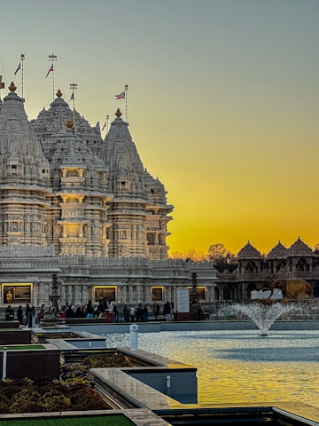 An intricately carved temple made from white stone stands against a backdrop of a warm, golden sky at sunset. The temple is adorned with decorative spires and flags. In front of the temple is a reflective water feature with a small fountain, surrounded by manicured gardens. People are gathered near the entrance, suggesting a place of worship or cultural significance.