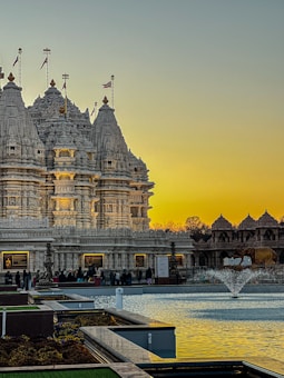 An intricately carved temple made from white stone stands against a backdrop of a warm, golden sky at sunset. The temple is adorned with decorative spires and flags. In front of the temple is a reflective water feature with a small fountain, surrounded by manicured gardens. People are gathered near the entrance, suggesting a place of worship or cultural significance.