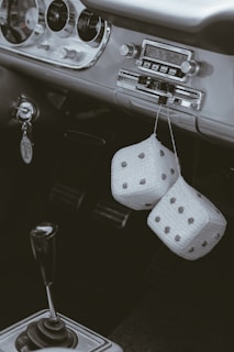 A vintage car interior featuring a classic dashboard with an old-fashioned radio and a manual gear shift. Two fuzzy dice dangle from the rearview mirror, adding a retro touch. The keychain has the brand name 'Ford' visible.