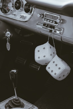 A vintage car interior featuring a classic dashboard with an old-fashioned radio and a manual gear shift. Two fuzzy dice dangle from the rearview mirror, adding a retro touch. The keychain has the brand name 'Ford' visible.