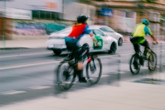 Two cyclists wearing helmets and bright vests ride along a city street, while a white car with green accents drives past them. The image is slightly blurred, indicating motion. The urban backdrop includes graffiti-covered walls and a building, contributing to a busy, lively atmosphere.