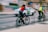 Two cyclists wearing helmets and bright vests ride along a city street, while a white car with green accents drives past them. The image is slightly blurred, indicating motion. The urban backdrop includes graffiti-covered walls and a building, contributing to a busy, lively atmosphere.