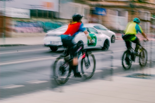 Two cyclists wearing helmets and bright vests ride along a city street, while a white car with green accents drives past them. The image is slightly blurred, indicating motion. The urban backdrop includes graffiti-covered walls and a building, contributing to a busy, lively atmosphere.