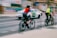 Two cyclists wearing helmets and bright vests ride along a city street, while a white car with green accents drives past them. The image is slightly blurred, indicating motion. The urban backdrop includes graffiti-covered walls and a building, contributing to a busy, lively atmosphere.