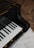 A close-up view of a sleek, black grand piano keyboard. The keys are glossy and well-polished, reflecting light beautifully, with a background of a wooden floor and a subtle rug.