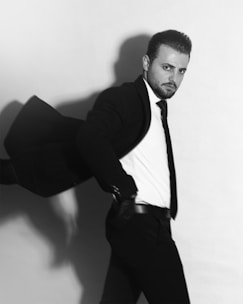A man in a suit appears to be in motion, as if turning quickly. He has a serious expression and is dressed in a dark blazer, white shirt, and tie. The background is plain, and the image is in black and white, contributing to a dramatic and professional atmosphere.