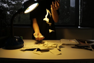 A person stands at a desk under a lamp, holding and tossing pieces of paper into the air. The scattered papers create a chaotic scene. The dim lighting and the glow from the lamp cast dramatic shadows.