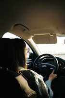 A person is seated in the driver's seat of a car, holding the steering wheel with one hand. They have long hair and are wearing sunglasses and a striped shirt. Sunlight is coming through the car's windows.