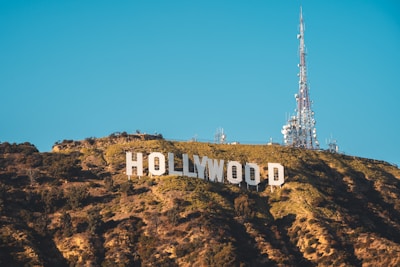 Large white letters spell out 'HOLLYWOOD' on a hillside covered with green foliage. A tall radio tower stands on the right side, reaching into a clear blue sky.
