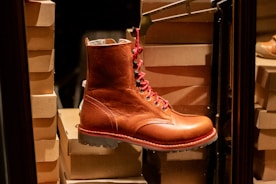 A single brown leather boot with red laces is prominently displayed against a backdrop of stacked cardboard shoe boxes. The boot has a rugged sole and detailed stitching, exuding a classic, sturdy appearance.