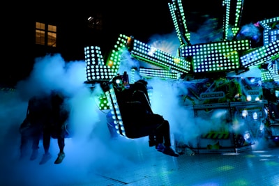 Brightly lit amusement ride with colorful LED lights surrounded by clouds of smoke, creating an energetic and futuristic atmosphere. The ride structure is covered with blue and green lights and people are visible in the seats, blurred by the smoke.