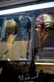Three ornately designed Venetian masks are displayed in a shop window. The first mask is gold with round eye accents, the second is a sleek black with minimalist features, and the third is an intricate red and gold design with elaborate swirls and patterns. The masks have elongated noses reminiscent of the traditional plague doctor style.