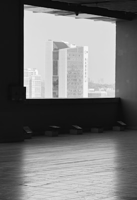 A cityscape is seen through a large, rectangular window, featuring a tall, modern skyscraper with vertical lines and glass windows. The floor leading to the window has a tiled pattern, and there is a contrast between the indoor shadows and the bright outdoor light. The scene is rendered in black and white, giving it a timeless, classic feel.