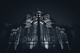 An ornate metal gate with intricate patterns and designs stands prominently, illuminated from behind by a light source that creates a dramatic contrast. The structure behind the gate seems to be a building with domed elements.