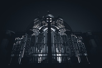 An ornate metal gate with intricate patterns and designs stands prominently, illuminated from behind by a light source that creates a dramatic contrast. The structure behind the gate seems to be a building with domed elements.