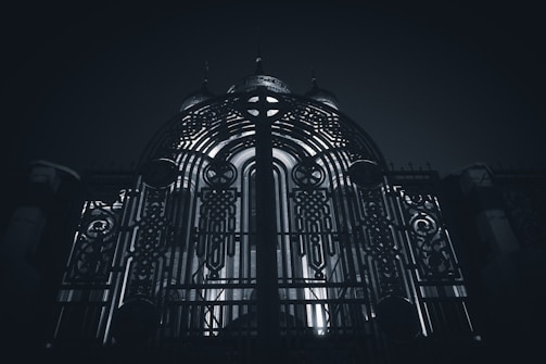 An ornate metal gate with intricate patterns and designs stands prominently, illuminated from behind by a light source that creates a dramatic contrast. The structure behind the gate seems to be a building with domed elements.