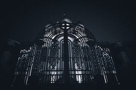 An ornate metal gate with intricate patterns and designs stands prominently, illuminated from behind by a light source that creates a dramatic contrast. The structure behind the gate seems to be a building with domed elements.