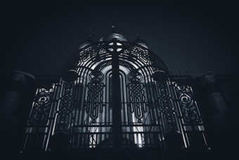 An ornate metal gate with intricate patterns and designs stands prominently, illuminated from behind by a light source that creates a dramatic contrast. The structure behind the gate seems to be a building with domed elements.