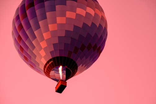 A hot air balloon with a colorful checkered pattern, primarily in shades of purple and red, floats against a backdrop of a pink sky. The flame inside the balloon's burner is visible, illuminating the balloon's interior.
