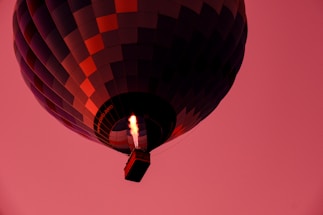 A hot air balloon with a patchwork pattern of dark blue and red squares floats against a bright pink sky. The flames that heat the air inside the balloon are prominently visible beneath the balloon's envelope, casting a warm glow.