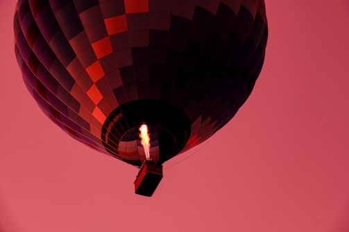 A hot air balloon with a patchwork pattern of dark blue and red squares floats against a bright pink sky. The flames that heat the air inside the balloon are prominently visible beneath the balloon's envelope, casting a warm glow.