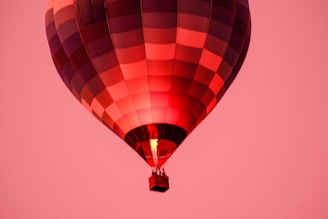 A hot air balloon floats against a vibrant pink sky. The balloon's canopy displays a patchwork of red and purple hues, and a flame is visible inside, providing the lift. Below, a small basket holds passengers.