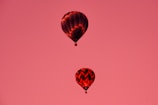 Two hot air balloons float against a backdrop of a solid pink sky. The balloons feature different patterns, with one displaying a checkerboard design and the other a chevron pattern. The lighting conveys a warm glow, enhancing the balloons' red and black colors.