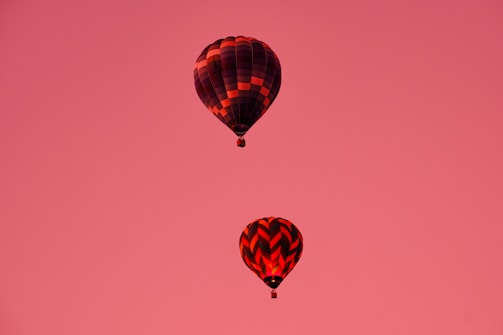 Two hot air balloons float against a backdrop of a solid pink sky. The balloons feature different patterns, with one displaying a checkerboard design and the other a chevron pattern. The lighting conveys a warm glow, enhancing the balloons' red and black colors.
