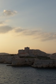 A historic fortress stands atop a rocky shoreline as the sun sets, casting a warm glow over the landscape. The sea is calm, with gentle waves lapping against the cliffs. The sky is a mix of soft blues and oranges, scattered with a few clouds.