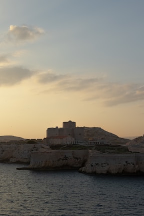 A historic fortress stands atop a rocky shoreline as the sun sets, casting a warm glow over the landscape. The sea is calm, with gentle waves lapping against the cliffs. The sky is a mix of soft blues and oranges, scattered with a few clouds.