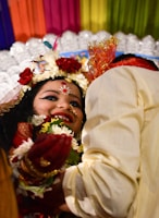 A joyous scene featuring two individuals, with one prominently wearing festive traditional attire. The person at the forefront has intricate facial makeup, floral decorations, and jewelry, all of which denote festivity and celebration. Colorful drapes in the background and a bright, happy demeanor enhance the lively atmosphere. The second individual is partially visible, wearing light-colored clothing and possibly interacting with the first person.