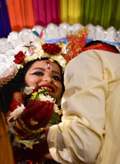 A joyous scene featuring two individuals, with one prominently wearing festive traditional attire. The person at the forefront has intricate facial makeup, floral decorations, and jewelry, all of which denote festivity and celebration. Colorful drapes in the background and a bright, happy demeanor enhance the lively atmosphere. The second individual is partially visible, wearing light-colored clothing and possibly interacting with the first person.