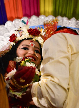 A joyous scene featuring two individuals, with one prominently wearing festive traditional attire. The person at the forefront has intricate facial makeup, floral decorations, and jewelry, all of which denote festivity and celebration. Colorful drapes in the background and a bright, happy demeanor enhance the lively atmosphere. The second individual is partially visible, wearing light-colored clothing and possibly interacting with the first person.