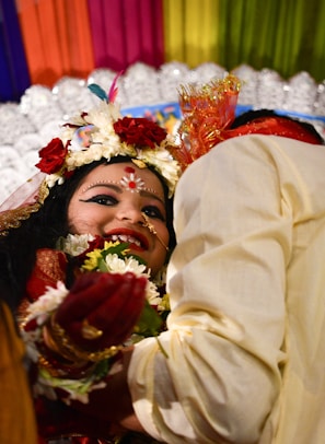 A joyous scene featuring two individuals, with one prominently wearing festive traditional attire. The person at the forefront has intricate facial makeup, floral decorations, and jewelry, all of which denote festivity and celebration. Colorful drapes in the background and a bright, happy demeanor enhance the lively atmosphere. The second individual is partially visible, wearing light-colored clothing and possibly interacting with the first person.