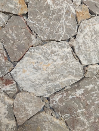 Rough stone tiles are laid out in an uneven pattern. The surfaces feature white and beige veins running across various shades of gray and brown. The texture appears rugged and natural, with some stones having sharper edges, while others seem more rounded.