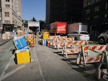 A busy city street scene with moving and delivery trucks parked alongside, surrounded by various equipment and boxes. Traffic cones and barricades labeled 'DANELLA' line the roadway. Several vehicles, including a white car and a red moving truck, are visible. Pedestrians and large buildings flank the street.