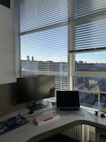 A workspace setup on a white desk near a large window with blinds partially drawn. The desk contains a monitor, a laptop that is open, a white keyboard with red and white keys, a mouse, and some miscellaneous items including a pair of glasses.