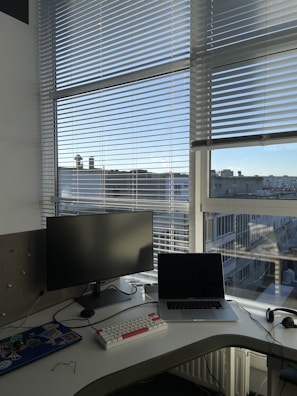 A workspace setup on a white desk near a large window with blinds partially drawn. The desk contains a monitor, a laptop that is open, a white keyboard with red and white keys, a mouse, and some miscellaneous items including a pair of glasses.