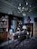 An elegant study room with an antique wooden desk and a vintage globe. The room includes a large wooden bookshelf filled with books, a decorative rug on the floor, and a chandelier hanging from the ceiling. A bust sits atop a tall wooden dresser, and an ornate lamp is placed on the desk.