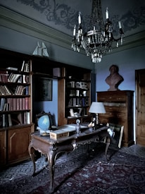 An elegant study room with an antique wooden desk and a vintage globe. The room includes a large wooden bookshelf filled with books, a decorative rug on the floor, and a chandelier hanging from the ceiling. A bust sits atop a tall wooden dresser, and an ornate lamp is placed on the desk.