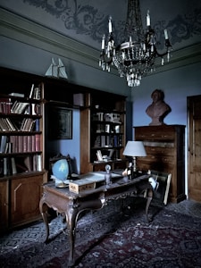 An elegant study room with an antique wooden desk and a vintage globe. The room includes a large wooden bookshelf filled with books, a decorative rug on the floor, and a chandelier hanging from the ceiling. A bust sits atop a tall wooden dresser, and an ornate lamp is placed on the desk.