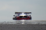 A distorted rear view of a racing car on an asphalt surface, creating a mirage effect with heat waves emanating from the road. The car is low and wide with a prominent rear spoiler.