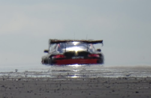 A distorted rear view of a racing car on an asphalt surface, creating a mirage effect with heat waves emanating from the road. The car is low and wide with a prominent rear spoiler.