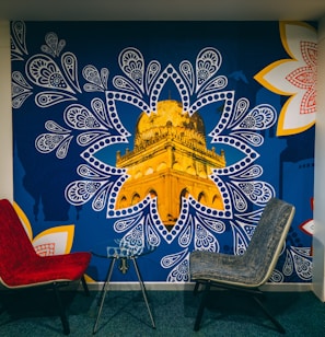 A mural depicting a golden architectural structure, prominently showcased within a blue, paisley-inspired pattern. The mural is accented by large, intricate white and red floral designs on the corners. In front of the mural, there are two modern chairs, one red and one gray, positioned around a small glass table.