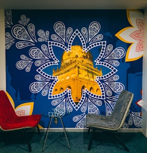 A mural depicting a golden architectural structure, prominently showcased within a blue, paisley-inspired pattern. The mural is accented by large, intricate white and red floral designs on the corners. In front of the mural, there are two modern chairs, one red and one gray, positioned around a small glass table.