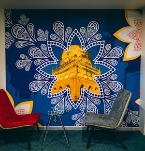 A mural depicting a golden architectural structure, prominently showcased within a blue, paisley-inspired pattern. The mural is accented by large, intricate white and red floral designs on the corners. In front of the mural, there are two modern chairs, one red and one gray, positioned around a small glass table.