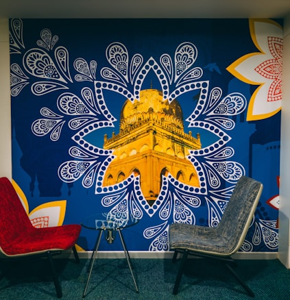 A mural depicting a golden architectural structure, prominently showcased within a blue, paisley-inspired pattern. The mural is accented by large, intricate white and red floral designs on the corners. In front of the mural, there are two modern chairs, one red and one gray, positioned around a small glass table.