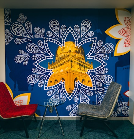 A mural depicting a golden architectural structure, prominently showcased within a blue, paisley-inspired pattern. The mural is accented by large, intricate white and red floral designs on the corners. In front of the mural, there are two modern chairs, one red and one gray, positioned around a small glass table.