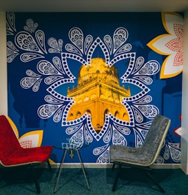 A mural depicting a golden architectural structure, prominently showcased within a blue, paisley-inspired pattern. The mural is accented by large, intricate white and red floral designs on the corners. In front of the mural, there are two modern chairs, one red and one gray, positioned around a small glass table.