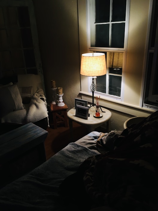 A cozy indoor setting features a warmly lit lamp on a small round table next to a window. The soft light casts gentle shadows across the room, highlighting a sofa with cushions and a knit blanket. Nearby, candles and decorative items add to the inviting atmosphere.